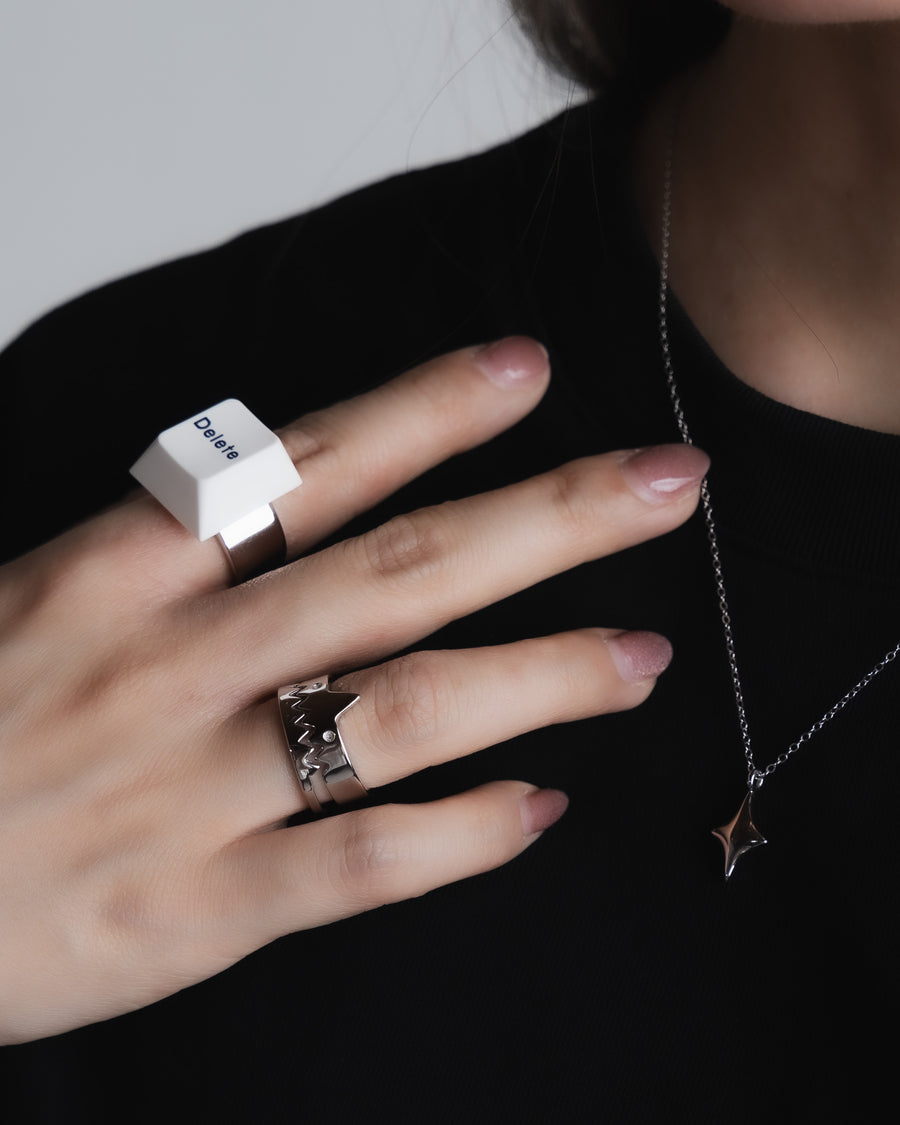 Hand shown against a black background, adorned with a unique keycap ring on the middle finger, characterized by its large, square shape with a prominent white keycap design. The ring finger showcases a creative monster design ring with a jagged, tooth-like pattern,  subtle star-shaped pendant on a silver necklace
