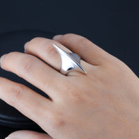 Close-up of a hand displaying a unique sterling silver ring with a bold, asymmetrical peak design. The ring's reflective surface is highlighted against her skin and the contrasting dark background