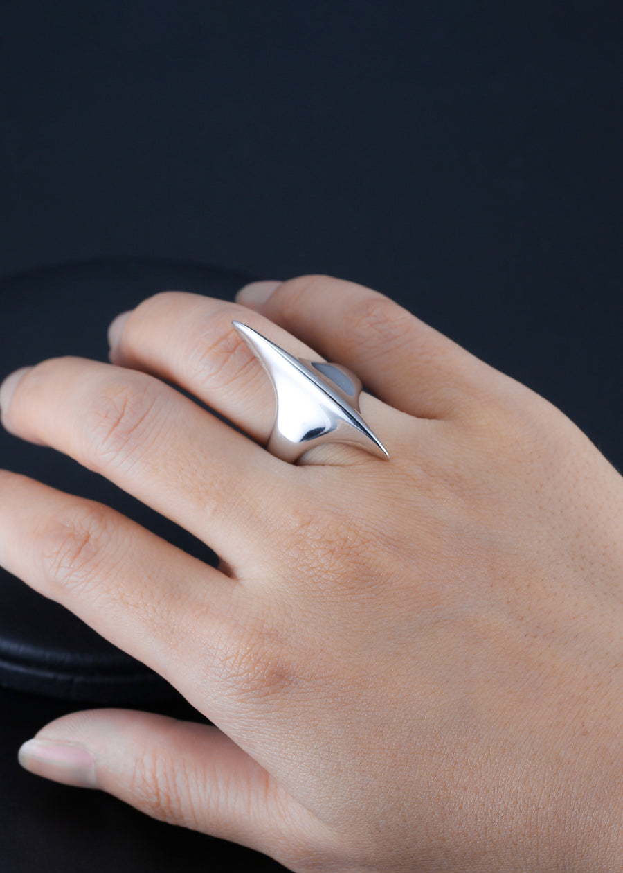 Close-up of a hand displaying a unique sterling silver ring with a bold, asymmetrical peak design. The ring's reflective surface is highlighted against her skin and the contrasting dark background